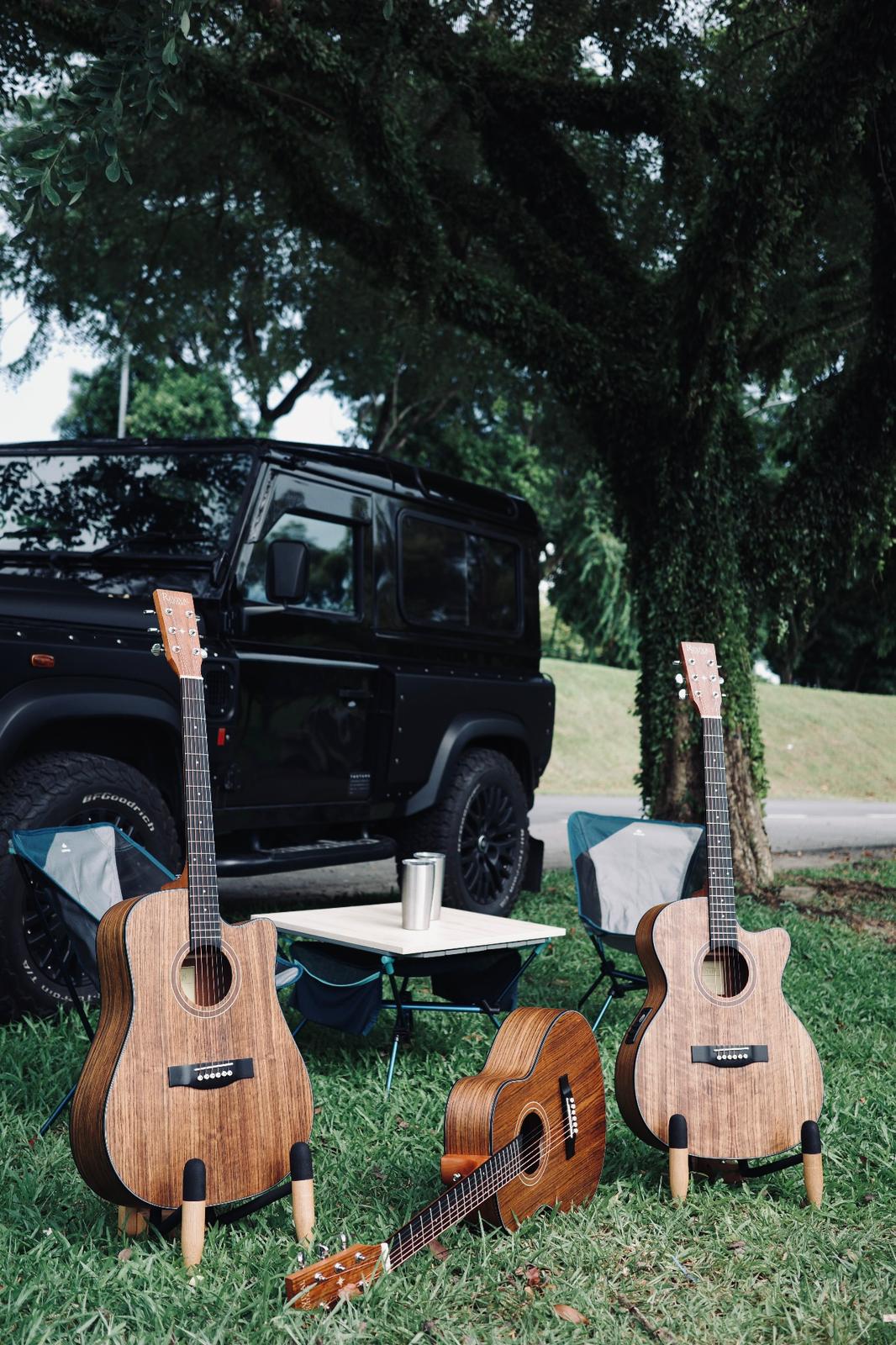 Three Revival Guitars walnut acoustics on the grass beside camp chairs and a Land Rover Defender